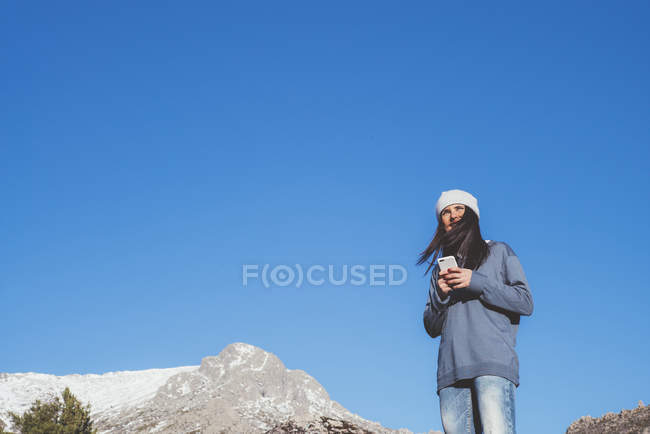 Frau mit Smartphone steht auf Felsen vor blauem Himmel — Stockfoto