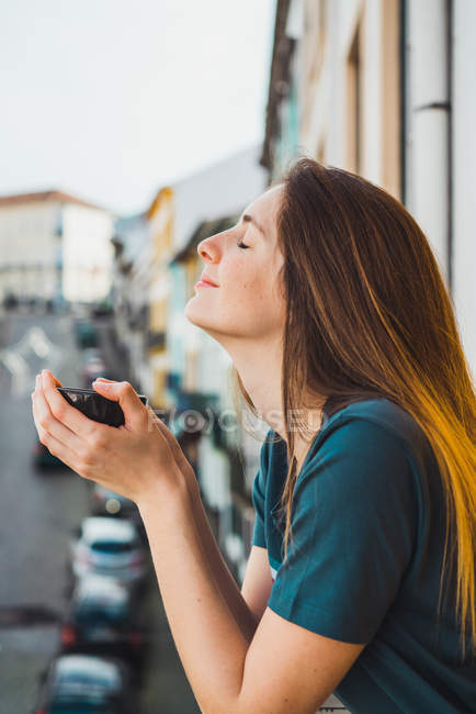 Morena sosteniendo la taza en las manos y posando con los ojos cerrados en el balcón - foto de stock