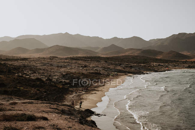 Distant view of people standing on coast with hills at seaside. — Stock Photo