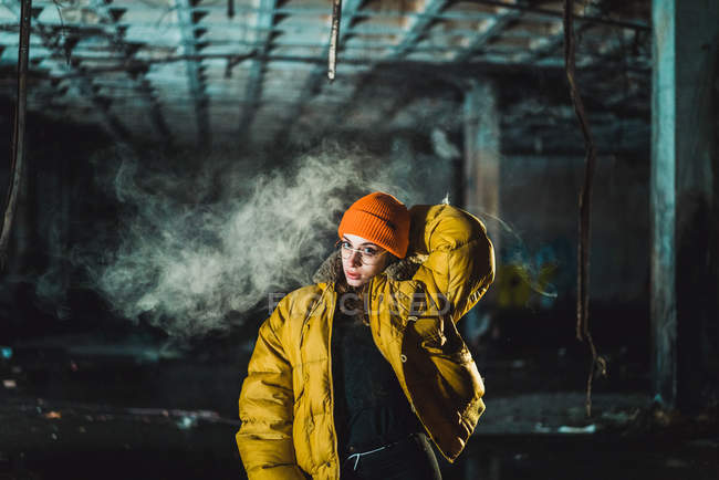 Молода жінка в жовтій куртці позує на тлі диму в покинутій будівлі — стокове фото
