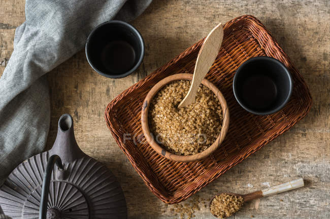 Directly above view of bowl full of brown sugar on wicker tray and teapot with cups — Stock Photo