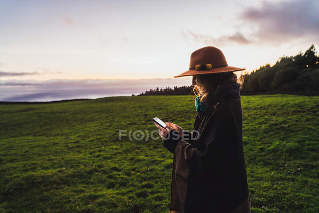 Vista lateral de la mujer en el teléfono inteligente de navegación sombrero en campos verdes - foto de stock
