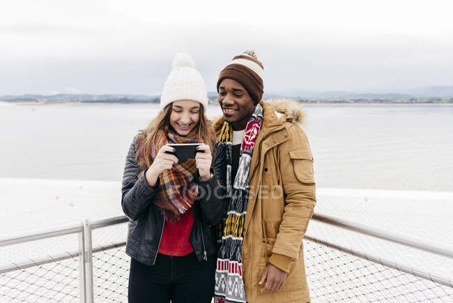 Весела пара, стоячи на паркані і дивлячись в смартфон проти океану . — стокове фото