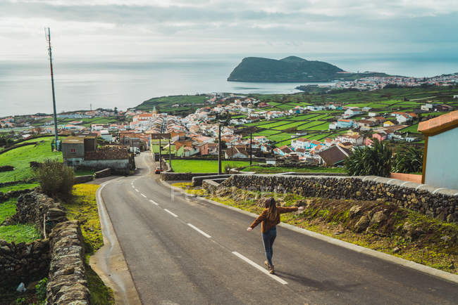 Mujer anónima con las manos separadas caminando por un camino vacío en una pequeña ciudad en la costa del océano . - foto de stock