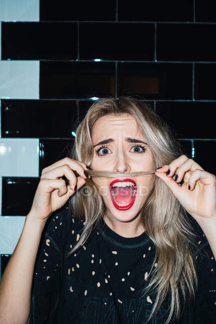 Blonde woman making mustache with hair and feeling scared at tiled wall. — Stock Photo