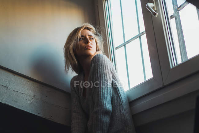 Mulher loira muito jovem sentada à janela e olhando para longe . — Fotografia de Stock