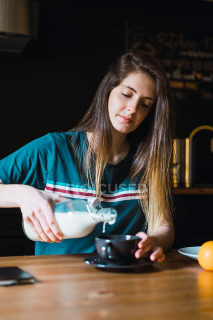 Femme brune assise à table et versant du lait dans une tasse — Photo de stock