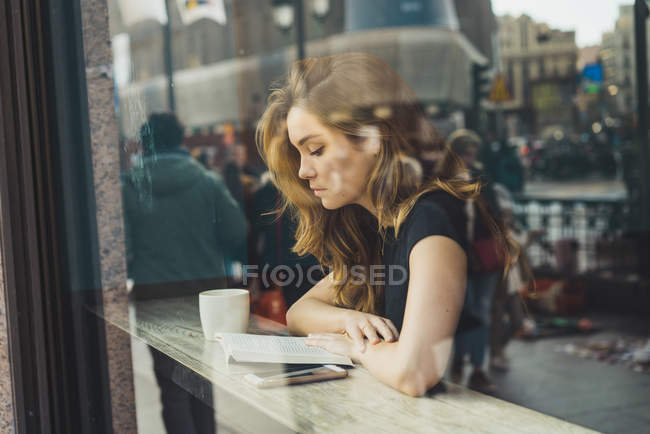 Vista a través de cristal de la joven leyendo libro en la cafetería - foto de stock