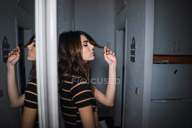 Side view of young woman leaning on wall and playing with hair in hall at home. — Stock Photo