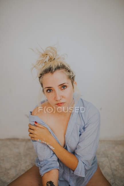 Donna attraente in camicia maschile seduta vicino al muro e guardando la fotocamera — Foto stock