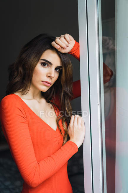Young woman in orange dress standing and looking at camera while leaning on window. — Stock Photo