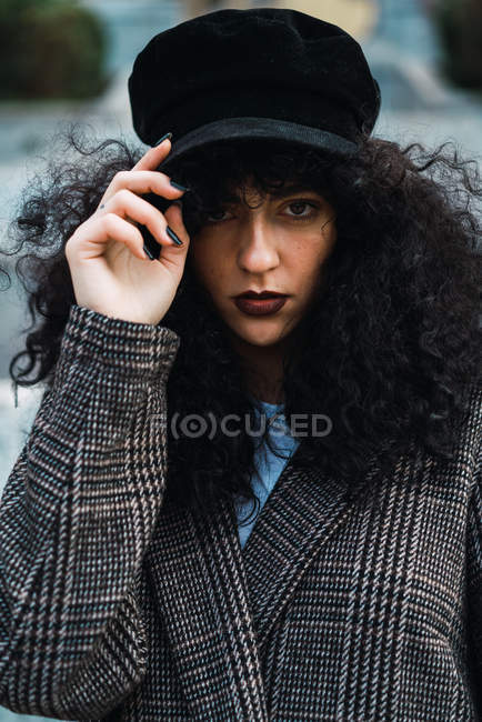 Portrait of stylish woman touching cap and looking at camera — Stock Photo