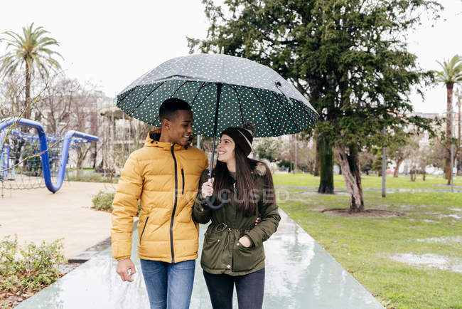 Cheerful couple under umbrella walking at park alley — Stock Photo