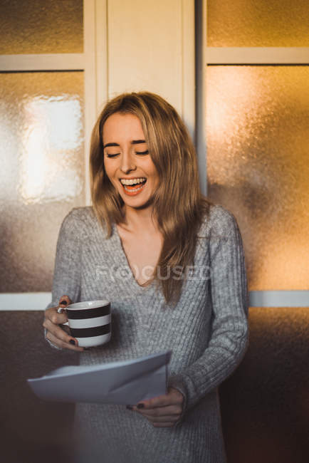 Сміється жінка з чашкою в руці дивиться на папери — стокове фото