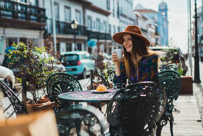 Smiling young woman in hat sitting with cup at cafe terrace. — Stock Photo
