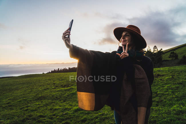 Cheerful woman in hat taking selfie at green fields — Stock Photo