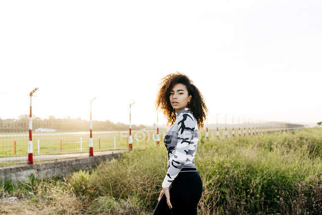Vista lateral da jovem com cabelo encaracolado olhando para a câmera e posando no campo . — Fotografia de Stock