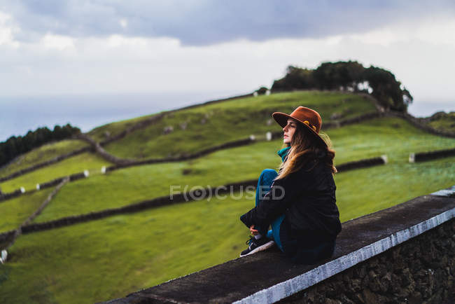 Young dreamy woman sitting on fence on background of green field on hill. — Stock Photo