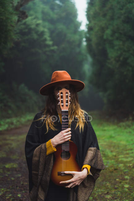 Charming woman hiding face with ukulele at road in woods — Stock Photo