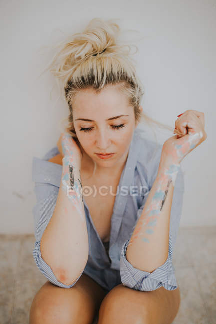 Attractive woman in male shirt and painted hands sitting by wall and looking down — Stock Photo