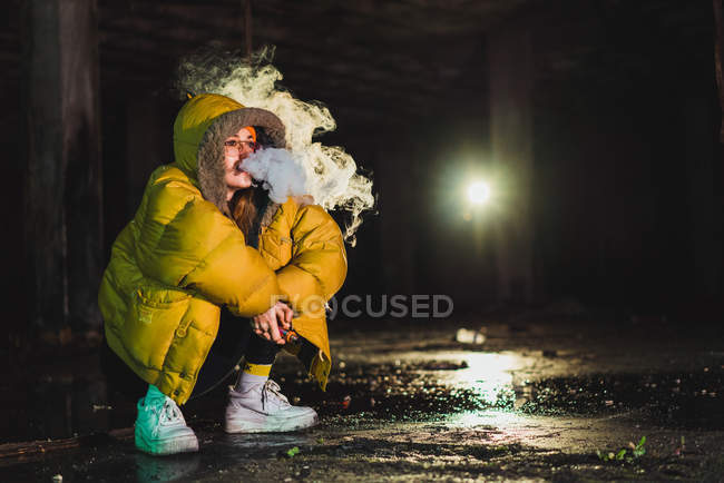 Jeune femme en veste assise et vaping dans un bâtiment abandonné . — Photo de stock