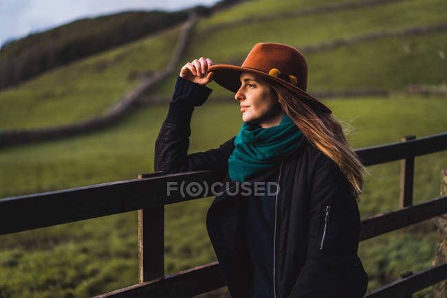 Мрійлива жінка в капелюсі стоїть на паркані і дивиться в сторону на зелене поле . — стокове фото