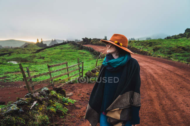 Woman in warm hat and scarf standing on empty countryside road at green fields — Stock Photo