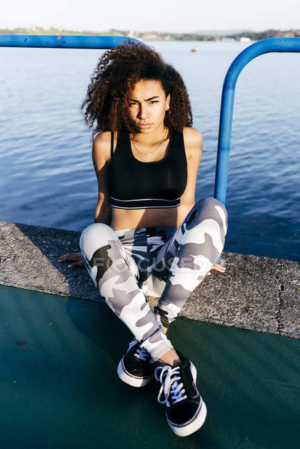 Young curly woman sitting on embankment at water and looking at camera. — Stock Photo