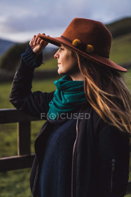 Dreamy woman standing at fence on field and touching hat — Stock Photo