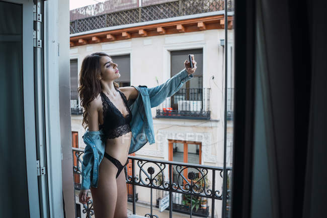 Vista lateral de mujer atractiva en lencería negra tomando selfie con smartphone en balcón . - foto de stock