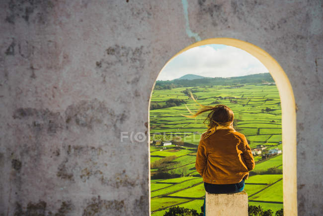 Rückansicht einer Frau, die im Bogen des Gebäudes auf grünen Feldern sitzt. — Stockfoto