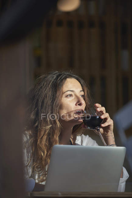 Mujer bebiendo vino mientras está sentado en la mesa con el ordenador portátil - foto de stock