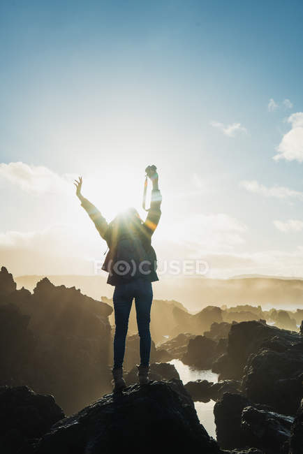 Vista posteriore della donna turista in posa con le braccia sollevate sulla scogliera nella giornata di sole . — Foto stock