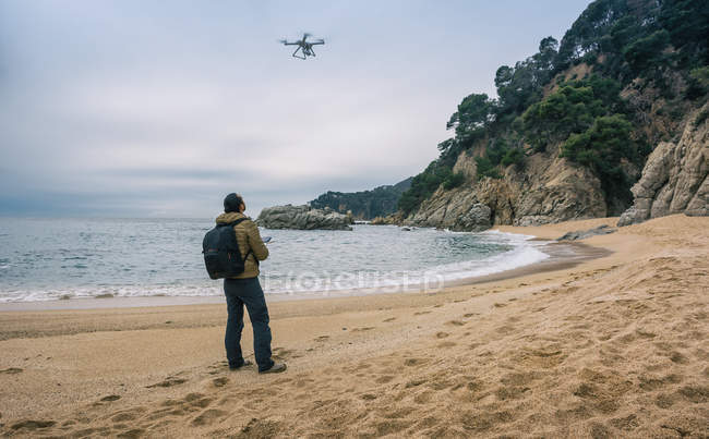 Vista posteriore dell'uomo con zaino in piedi sulla spiaggia e drone di prova in aria — Foto stock