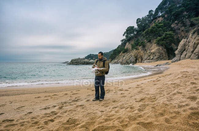 Чоловік з рюкзаком стоїть на пляжі і тестує безпілотник в повітрі — стокове фото