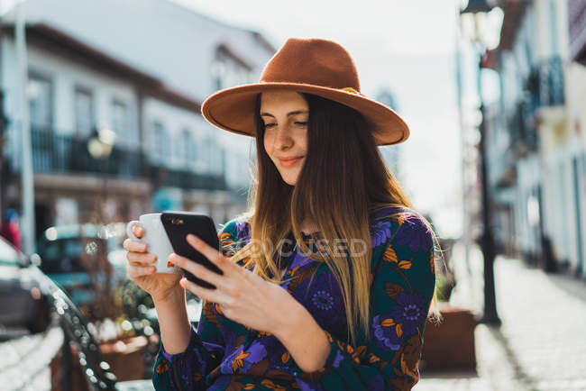 Женщина в шляпе сидит на террасе кафе с чашкой и смартфоном — стоковое фото