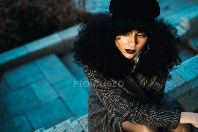 Young brunette curly woman in coat posing on fence at stairs in city park. — Stock Photo
