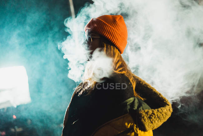 Side view of pretty young woman standing and vaping in back lit at night. — Stock Photo