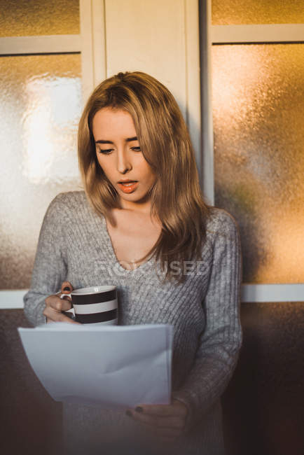 Jeune femme choquée avec tasse debout et des papiers de lecture à la maison . — Photo de stock