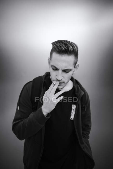 Porträt eines lässigen jungen Mannes, der vor grauem Hintergrund Zigarette raucht — Stockfoto