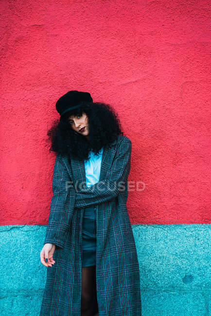 Молода брюнетка кучерява жінка стоїть в стильному пальто і позує на яскравій стіні . — стокове фото