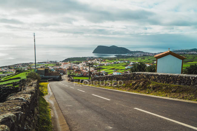 Landscape of running road among green valley to village on ocean coastline. — Stock Photo