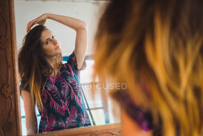 Jovem mulher de pé no espelho e olhando para a reflexão ao ajustar o cabelo . — Fotografia de Stock
