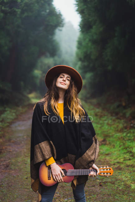 Charming woman posing with ukulele on rural road in woods — Stock Photo