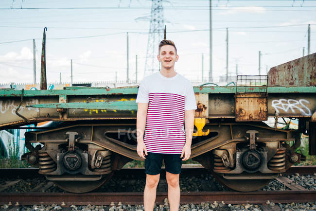Улыбающийся молодой человек, стоящий у угрюмого поезда и смотрящий в камеру — стоковое фото
