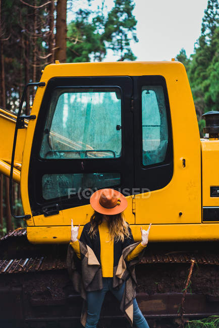 Stylish woman in hat and poncho showing rock and roll gesture on background of tractor. — Stock Photo