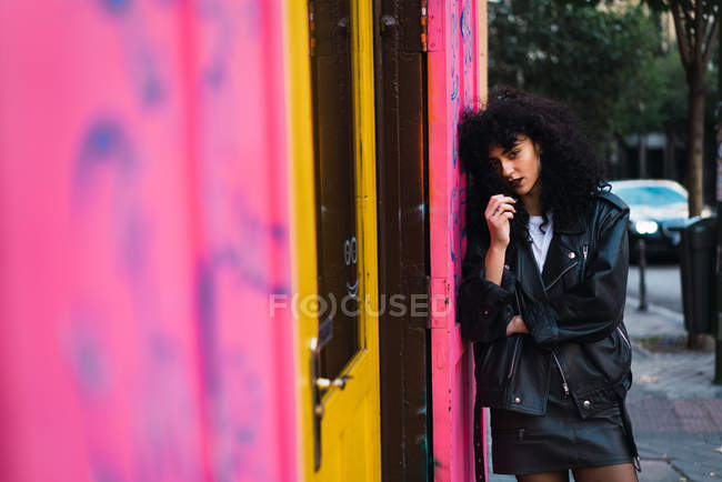 Glamour femme bouclée dans des vêtements en cuir appuyé sur le mur rose et regardant la caméra — Photo de stock