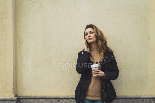 Femme posant avec smartphone et café à emporter au mur — Photo de stock