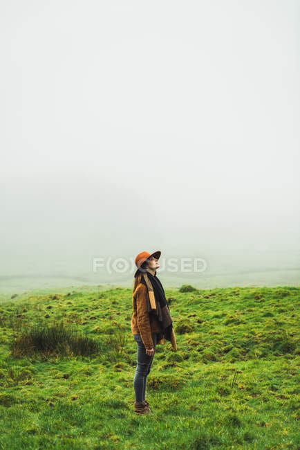 Vista laterale della donna in giacca calda in piedi e guardando verso l'alto in praterie verdi nebbiose . — Foto stock