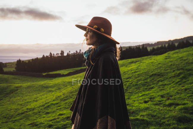 Vista lateral da mulher em chapéu elegante em pé no fundo de pastagens verdes e nuvens . — Fotografia de Stock
